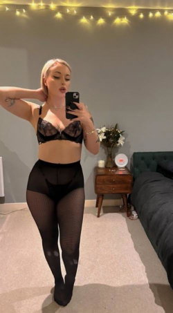 Blonde woman in black lace bra and fishnet stockings takes a mirror selfie in a cozy bedroom with string lights.