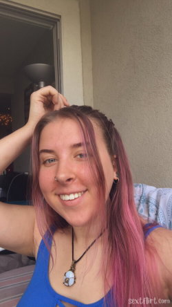 A woman with pink-tinted hair smiles at the camera, wearing a blue tank top and a pendant necklace, outdoors near a