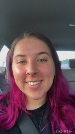 A woman with vibrant pink hair smiles in a car selfie, wearing a black top and seatbelt.