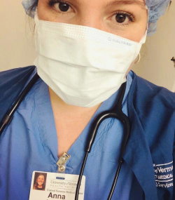 Woman in blue medical scrubs with stethoscope, wearing surgical mask and cap, ID badge visible, taking selfie.