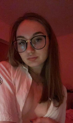 A woman with shoulder-length brown hair and glasses wears an unbuttoned white shirt, sitting in a dimly lit room with