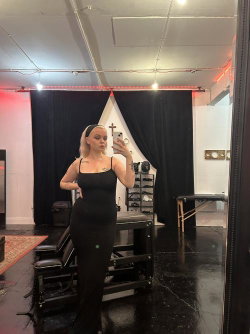 A blonde woman in a black sleeveless dress takes a mirror selfie in a dimly lit room with red accent lighting and black