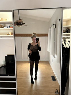 A woman with blonde hair in a bun takes a mirror selfie in a sheer black top, mini skirt, and black tights in a
