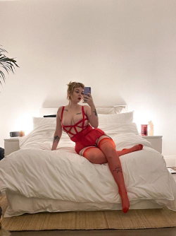 A woman with short blonde hair sits on a bed in red lingerie and thigh-high stockings, taking a mirror selfie with
