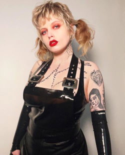 Blonde woman with pigtails and bold red lipstick wears a black latex outfit with buckles; multiple tattoos visible on