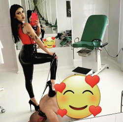 A woman with long dark hair and tattoos wears a red top and black leather pants, posing in a medical-style room while