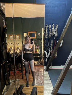 A woman in a black lace bra and high-waisted shorts poses in front of a mirror, wearing sheer black thigh-highs, in a