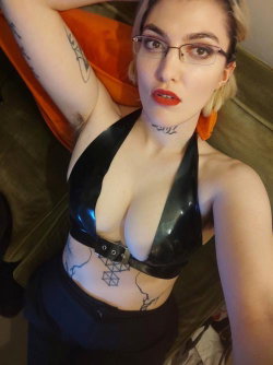 A woman with short blonde hair and glasses wears a black latex top, showing tattoos on her arm and torso, posing on a