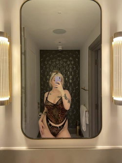 Blonde woman in leopard-print lingerie takes mirror selfie in a well-lit bathroom with patterned wall and sconce