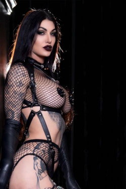 A woman with long dark hair and bold makeup wears a black fishnet bodysuit with leather harness and gloves, posing