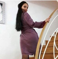 A woman with long dark hair in a form-fitting purple dress poses on a staircase, holding the railing and looking over