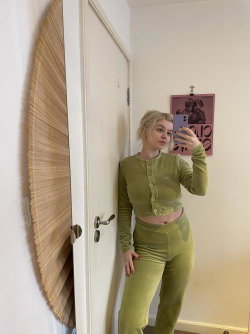Blonde woman in a matching olive green cropped cardigan and high-waisted pants takes a mirror selfie in a room with a