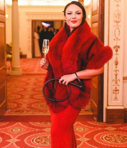 A woman in a red fur stole and matching dress holds a champagne flute in an ornate hallway.