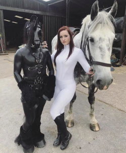 A woman in a white bodysuit and black boots stands beside a real horse and a person in a black horse costume, outdoors