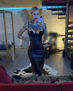 Blonde woman in a black leather dress with full arm tattoos takes a mirror selfie in a modern room with brick walls and