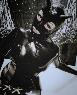 Woman in a shiny black latex catwoman costume with mask and horns, posing against a patterned backdrop.