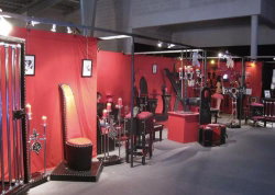 An indoor exhibition space with red walls, featuring various BDSM furniture and equipment including restraints, chairs,