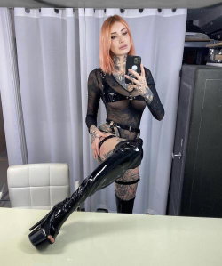 A woman with pink hair and extensive tattoos wears a black sheer top, harness, and shiny thigh-high boots while posing