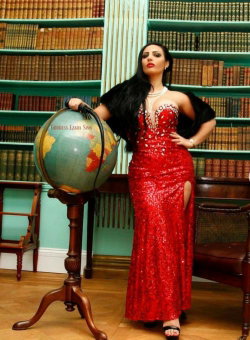 A woman with long dark hair in a red sequined gown poses beside a globe in a library with teal bookshelves.