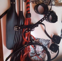 Assorted black and red leather BDSM accessories including whips, cuffs, and restraints arranged on a light surface.