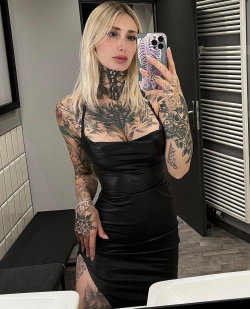 Blonde woman with extensive tattoos on arms, neck, and legs wearing a black leather dress, taking a mirror selfie in a