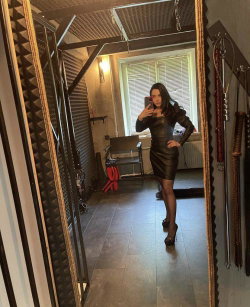 Brunette woman in a form-fitting black dress poses in a mirror selfie within a room featuring soundproofing panels and