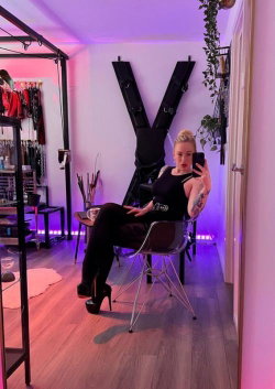 A woman with blonde hair and tattoos sits in a chair, taking a mirror selfie in a room with purple lighting, wearing
