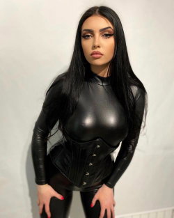 Woman with long black hair wearing a shiny black bodysuit and corset, posing against a plain white wall.