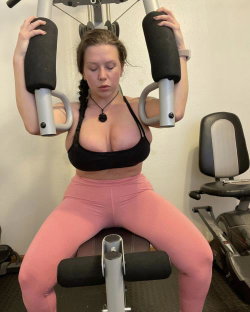 A woman with braided hair in a black sports bra and pink leggings performs a chest press exercise on gym equipment.
