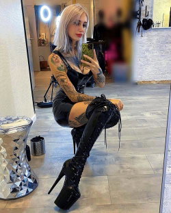 A blonde woman with tattoos takes a mirror selfie in a black latex outfit and tall platform boots, sitting on the floor