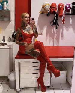 A blonde woman with extensive tattoos wears a red latex dress and thigh-high boots, sitting on a red-topped cabinet in