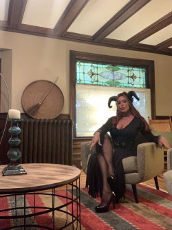A woman with long brown hair wearing a black horned costume sits in a chair in a room with stained glass and wooden