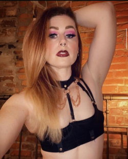 A woman with long auburn hair and bold makeup poses in a black crop top against a brick wall, one arm raised behind her