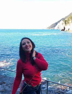 A woman with dark hair in a red turtleneck and jeans smiles by the ocean, standing near a railing with cliffs in the