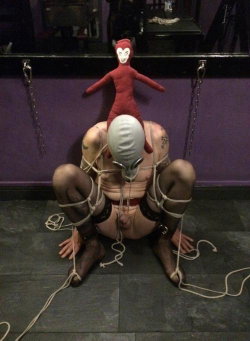 A person with a white mask and black stockings is bound with rope in a kneeling position, wearing a red thong, with a