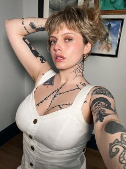 Blonde woman with tattoos on arms and chest, wearing a white button-up top, taking a selfie indoors.