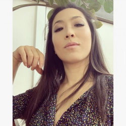 A woman with long dark hair and a patterned top poses in a selfie, looking directly at the camera with a neutral