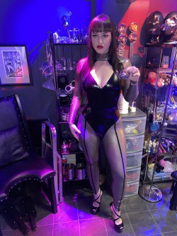 A woman in a shiny black bodysuit with silver arm sleeves and high heels poses in a dimly lit room with purple lighting