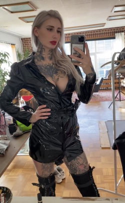Blonde woman with visible tattoos takes a mirror selfie in a black shiny outfit, wearing thigh-high boots in a bright