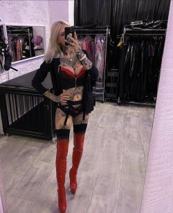 Blonde woman with full-body tattoos takes a mirror selfie in a red bustier, black harness, and thigh-high red boots in