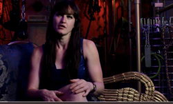 Brunette woman with long hair in a dark blue top, seated on a patterned couch in a dimly lit room with metallic decor.