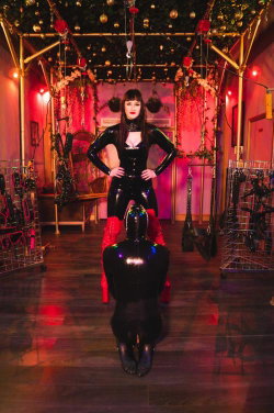A woman in a black latex bodysuit and red boots stands over a kneeling person in a dimly lit, decoratively adorned room