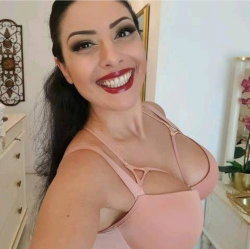 A smiling woman with long dark hair and red lipstick poses in a pink lingerie top, taking a selfie in a well-lit room
