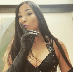 A woman with long dark hair wears a black leather corset and gloves, posing indoors with one hand near her lips.