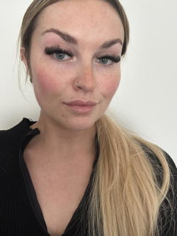 Blonde woman with freckles and makeup, wearing a black ribbed top, posing in a neutral indoor setting.