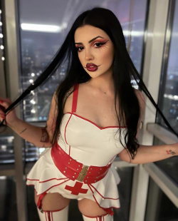 A woman with long dark hair and bold makeup wears a white and red nurse costume with thigh-high boots, posing indoors