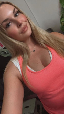 Blonde woman with long hair in a coral tank top, taking a close-up selfie indoors.