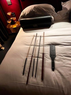 Several whips and a paddle arranged on a bed in a dimly lit room with drinks on a side table.