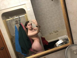 A woman with a curvy build takes a mirror selfie in a bathroom, nude, with towels and a bathtub visible in the