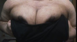 A muscular person with a thick build lifts their black shirt to reveal a hairy chest and large breasts.